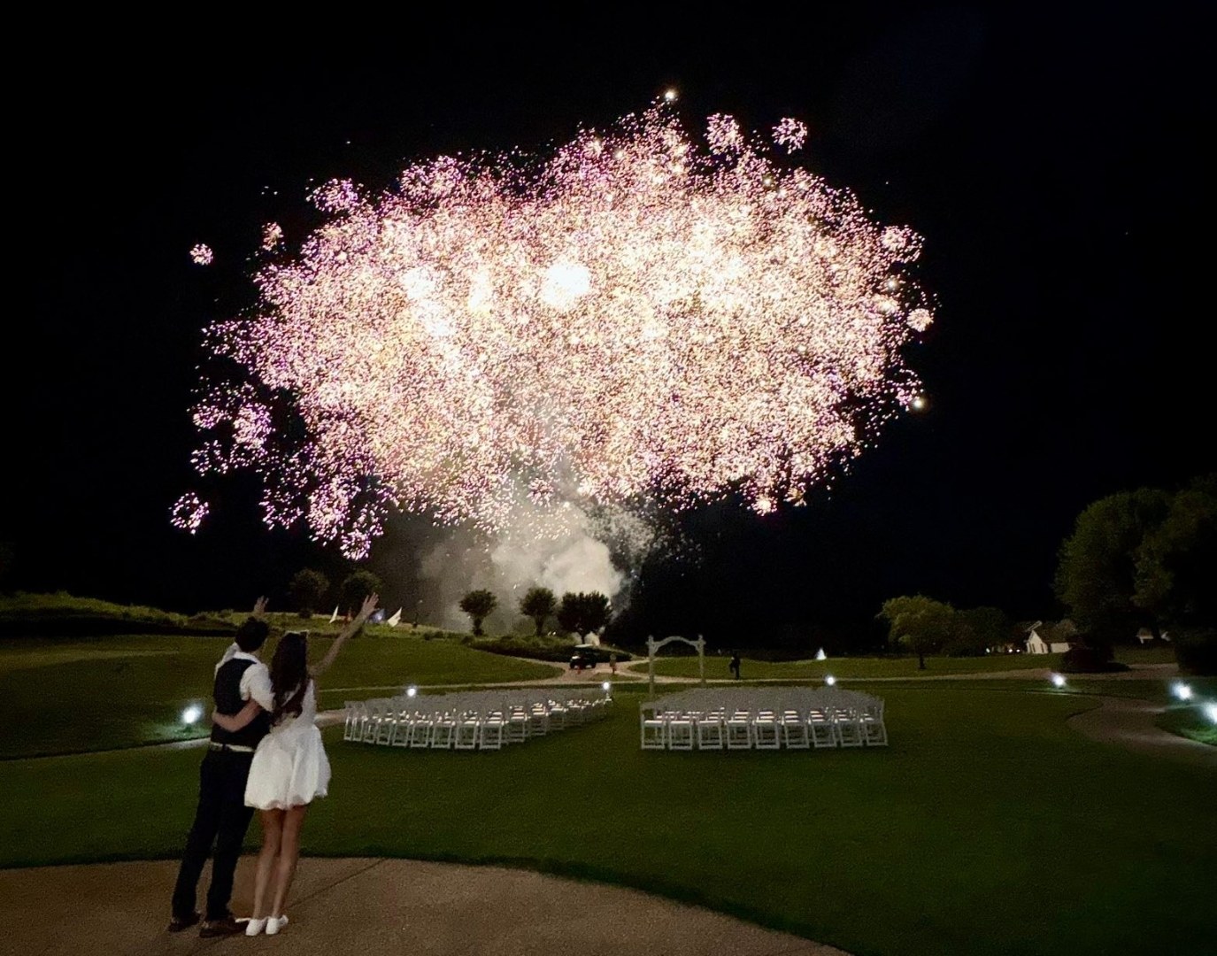 Bride and groom holding each other beneath a massive fireworks burst over their outdoor wedding ceremony